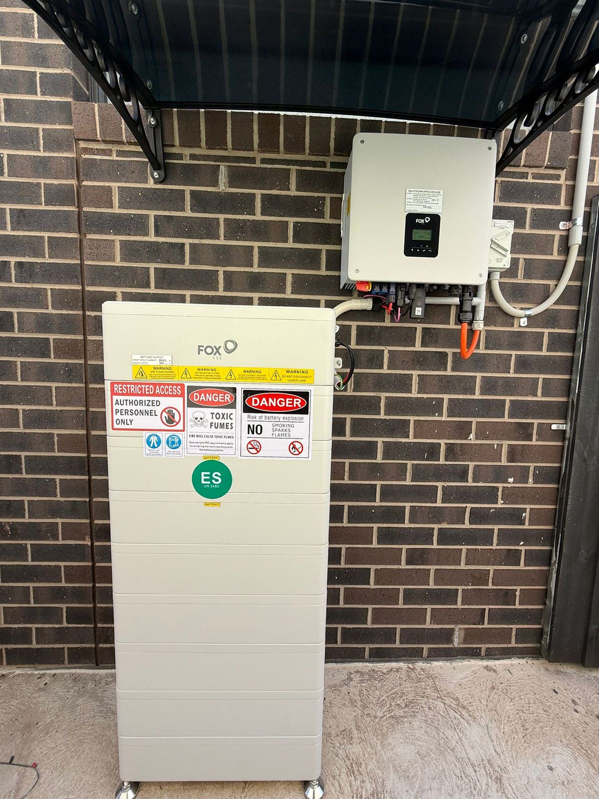 Outdoor Fox ESS battery and hybrid inverter installed on a brick wall under an awning