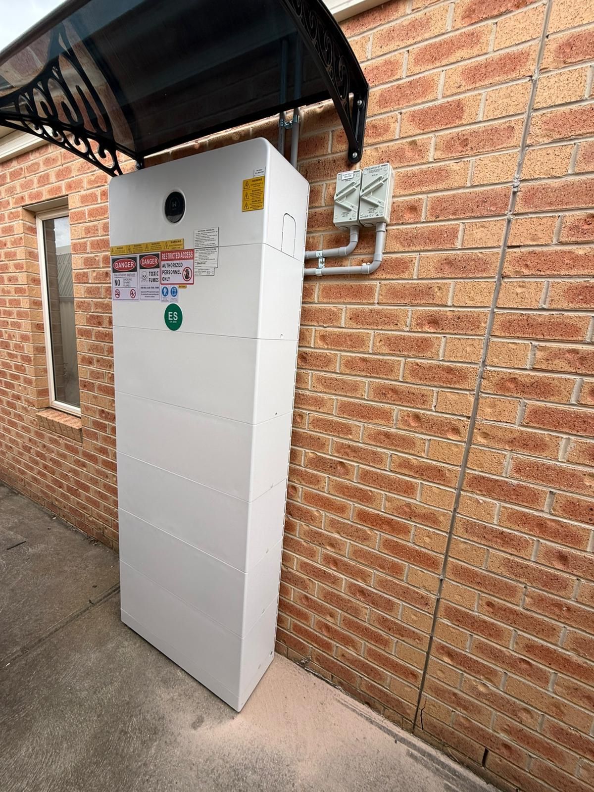 Tall outdoor battery stack installed on a brick wall under a window awning